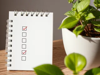 A spiral notepad with a ticked checklist beside a potted plant on a wooden table